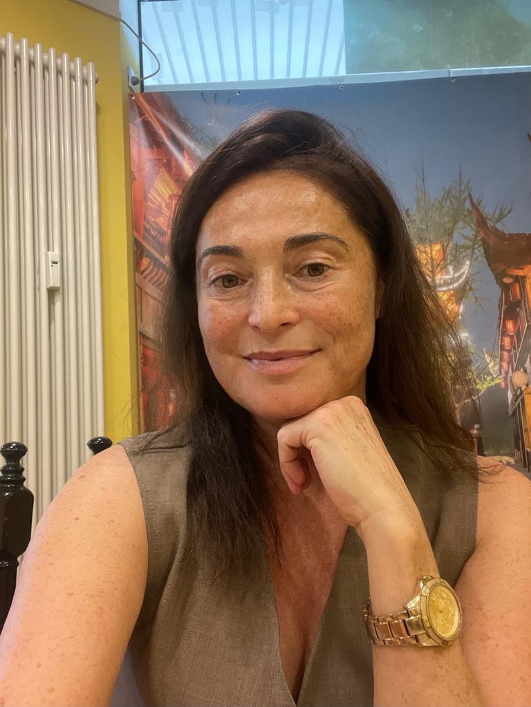 Woman with long dark hair wearing a brown sleeveless shirt and gold watch, resting her chin on her hand, smiling at camera indoors